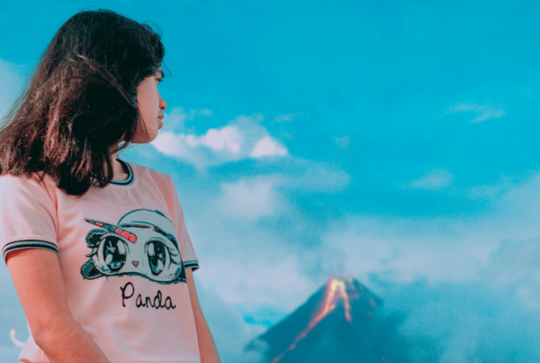 Una chica encarando la camara mira por encima del hombro detrás suya donde un volcan está en erupción.